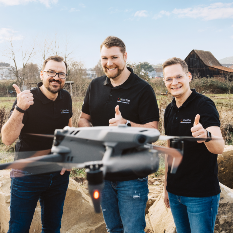 Drei lächelnde Mitarbeiter von Enerplan Energieberatung mit einem Quadrocopter draußen, zeigen Daumen hoch