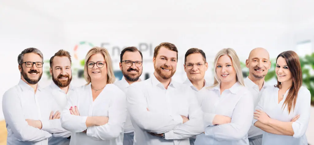 Teamfoto der Mitarbeiter von Enerplan Energieberatung, lächelnd und in weißen Hemden gekleidet, vor einem unscharfen Bürohintergrund mit dem Logo von Enerplan im Hintergrund.